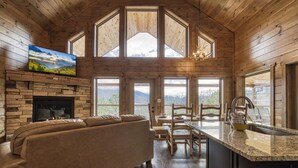 A cozy cabin interior with a fireplace, a sofa, a dining area, and large windows offering mountain views. - Smoky Mountain Dreamin'-Mountain Views, Hot Tub, $560 tickets each paid day! (Sevierville)