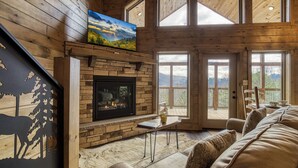 A cozy living room with a stone fireplace, a flat-screen TV mounted above it, and a large window offering a view of the outdoors. - Smoky Mountain Dreamin'-Mountain Views, Hot Tub, $560 tickets each paid day! (Sevierville)