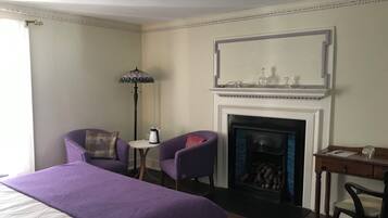 Une chambre d’hôtel avec un grand lit, deux fauteuils, une petite table avec une bouilloire, une cheminée et un bureau avec une chaise.