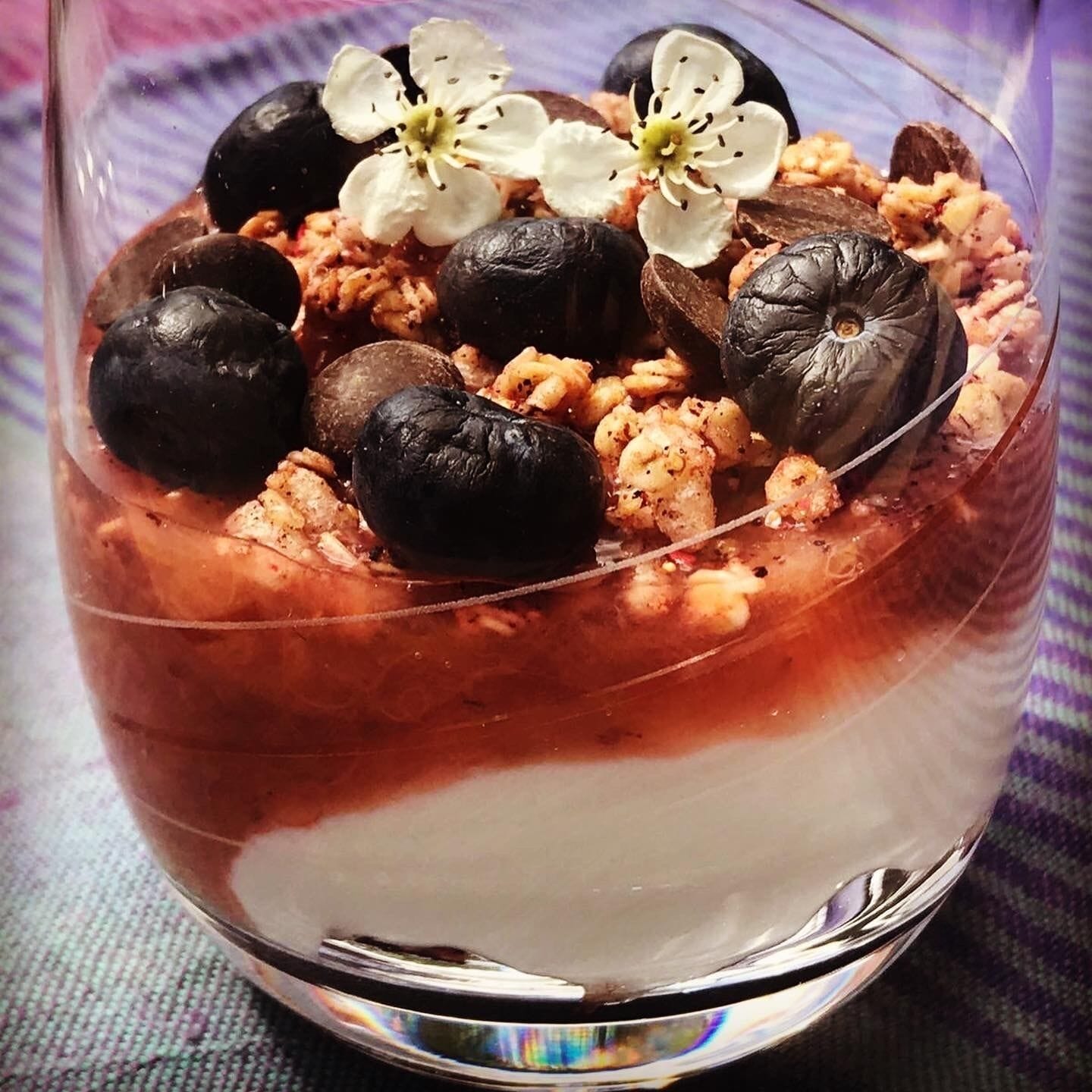 A dessert in a clear glass bowl with layers of chocolate and fruit, topped with black olives and white flowers.