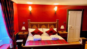 Un lit bien fait, recouvert d’une couverture à carreaux, avec deux lampes de chevet et un mur rouge agrémenté de deux appliques murales.