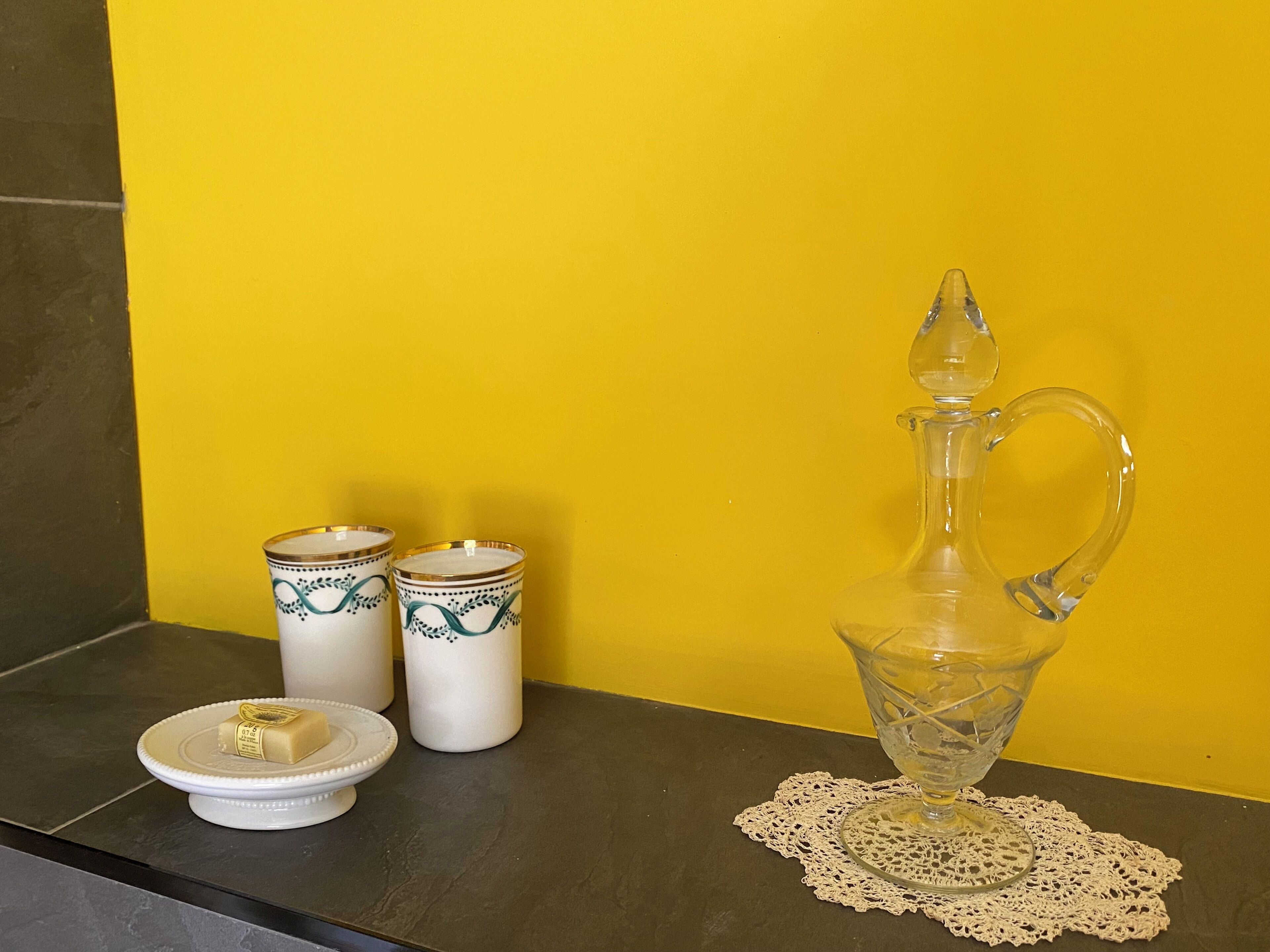 A kitchen counter with a clear glass pitcher, two white containers with green designs, and a small white plate with a bar of soap.