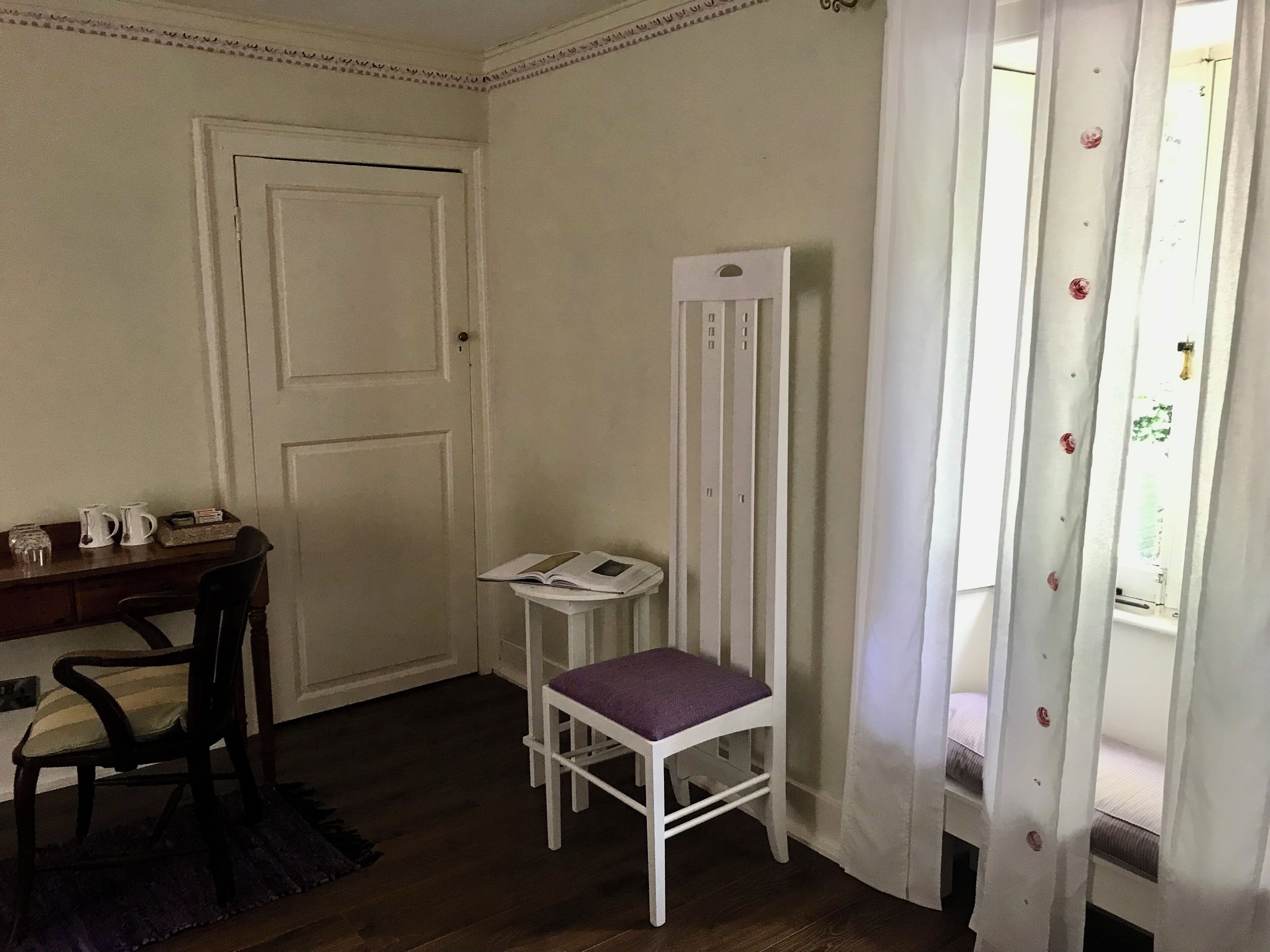 A room with a wooden desk, a white chair, a small white table, and a window with white curtains.