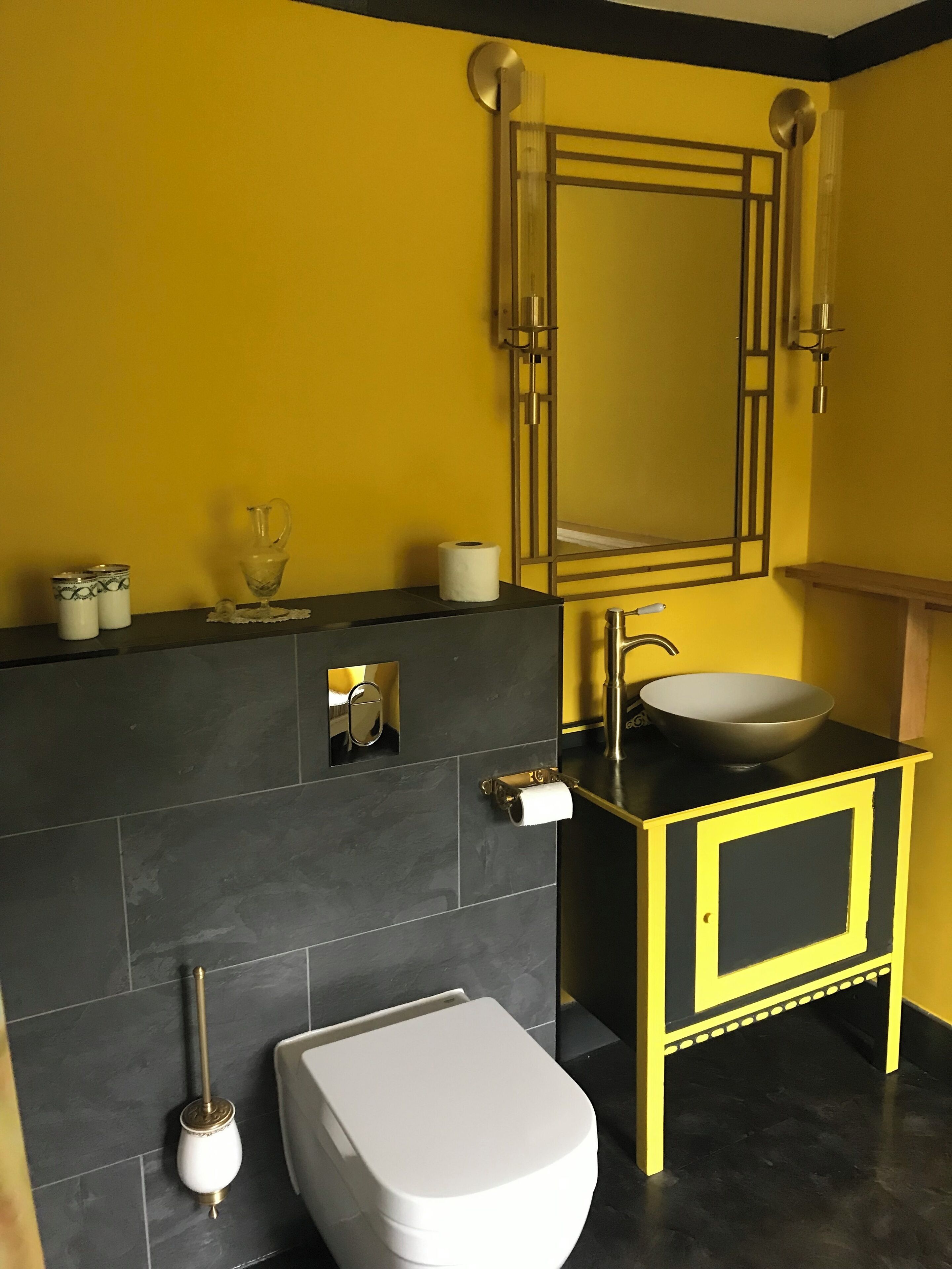 A bathroom with a yellow wall, a black and yellow vanity, a white toilet, and a mirror with a gold frame.