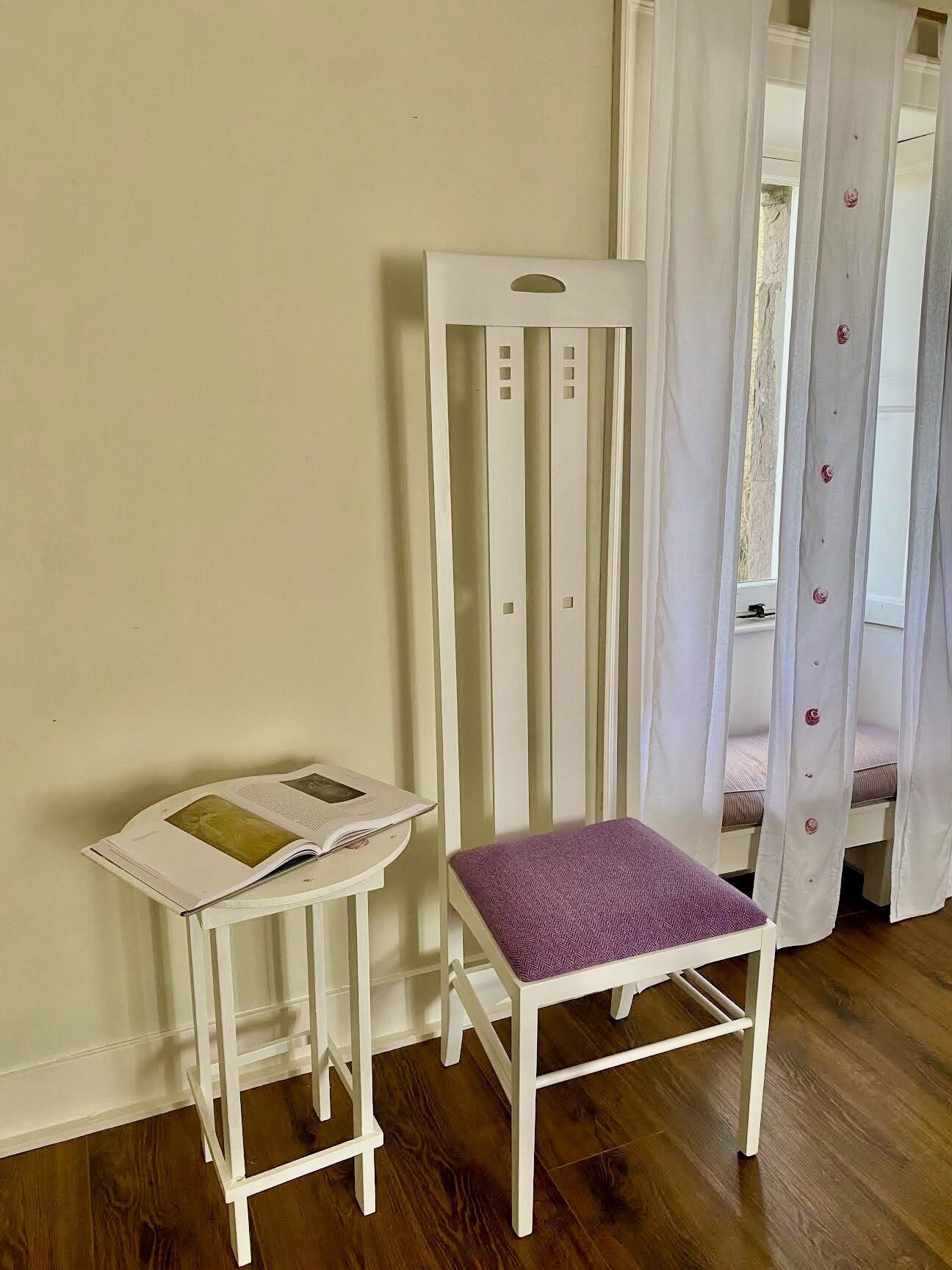 Une chaise en bois blanche avec un coussin violet, une petite table blanche sur laquelle se trouve un livre, et une fenêtre avec des rideaux blancs.