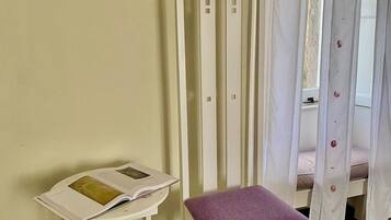 Une chaise en bois blanche avec un coussin violet, une petite table blanche sur laquelle se trouve un livre, et une fenêtre avec des rideaux blancs.