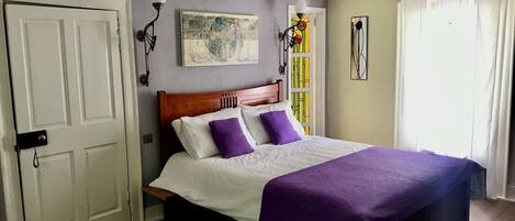 A bedroom with a wooden bed, white bedding, and a purple throw blanket. There is a door on the left and a window on the right.