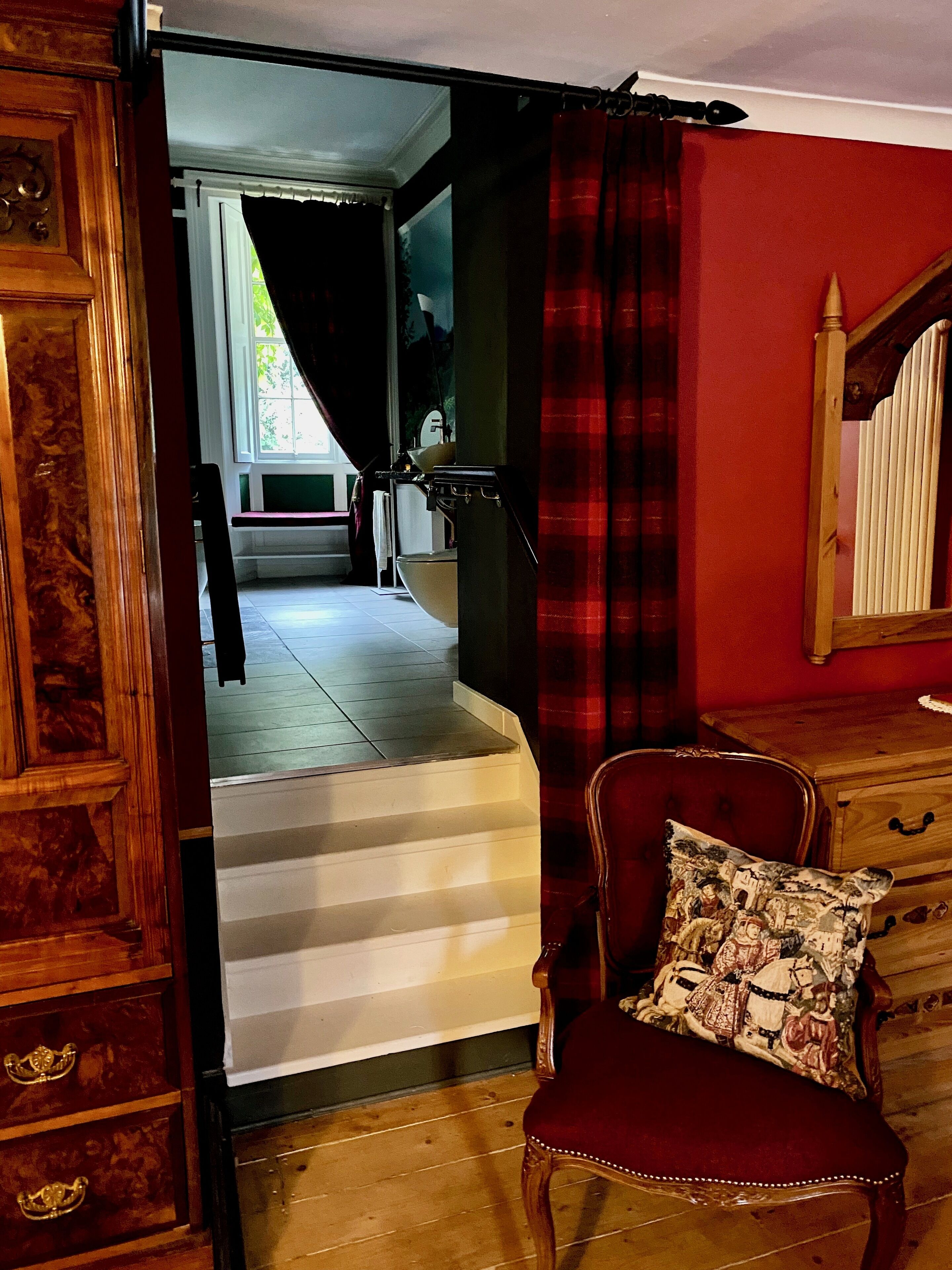 Quarto com cadeira de madeira, parede vermelha, escada e vista para um banheiro.