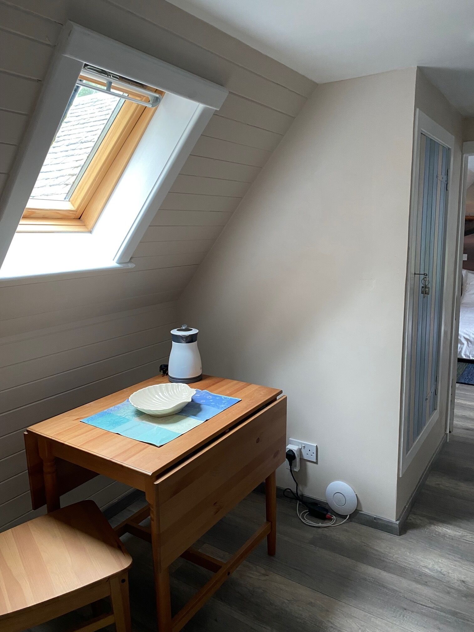 Ett litet, ombonat rum med ett sluttande tak, ett träbord med en vattenkokare, en skål och en matta. Det finns ett fönster med utsikt över omgivningen och en dörr som leder till ett annat rum.