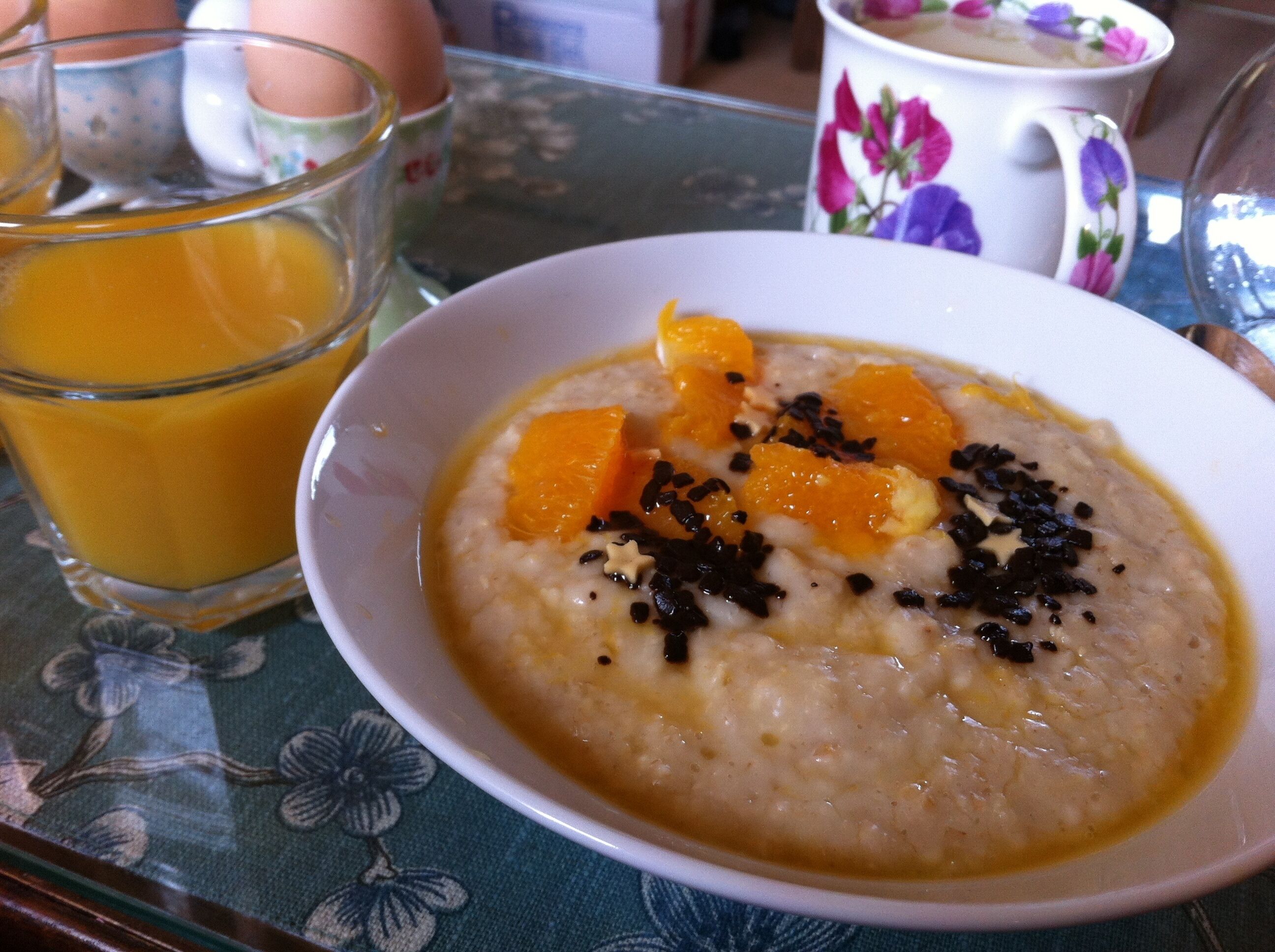 A bowl of oatmeal with orange pieces and black seeds, a glass of orange juice, and a floral mug on a table.