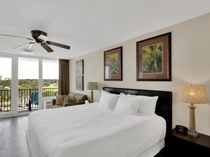 A hotel room with a large bed, a ceiling fan, a balcony with a view of the city, and two framed pictures on the wall.