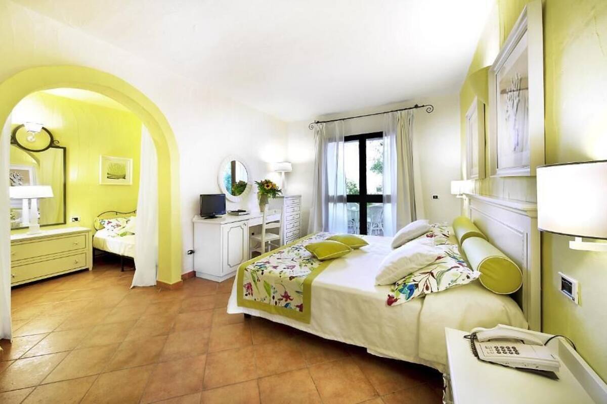 A hotel room with a bed, a desk, a chair, a lamp, a mirror, and a window with curtains.