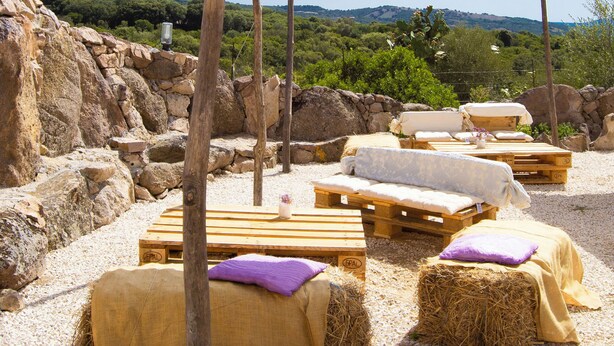 Outdoor seating with hay bales and wooden benches, a white canopy, and a rocky backdrop. - Pasare Glamping (Perfugas)