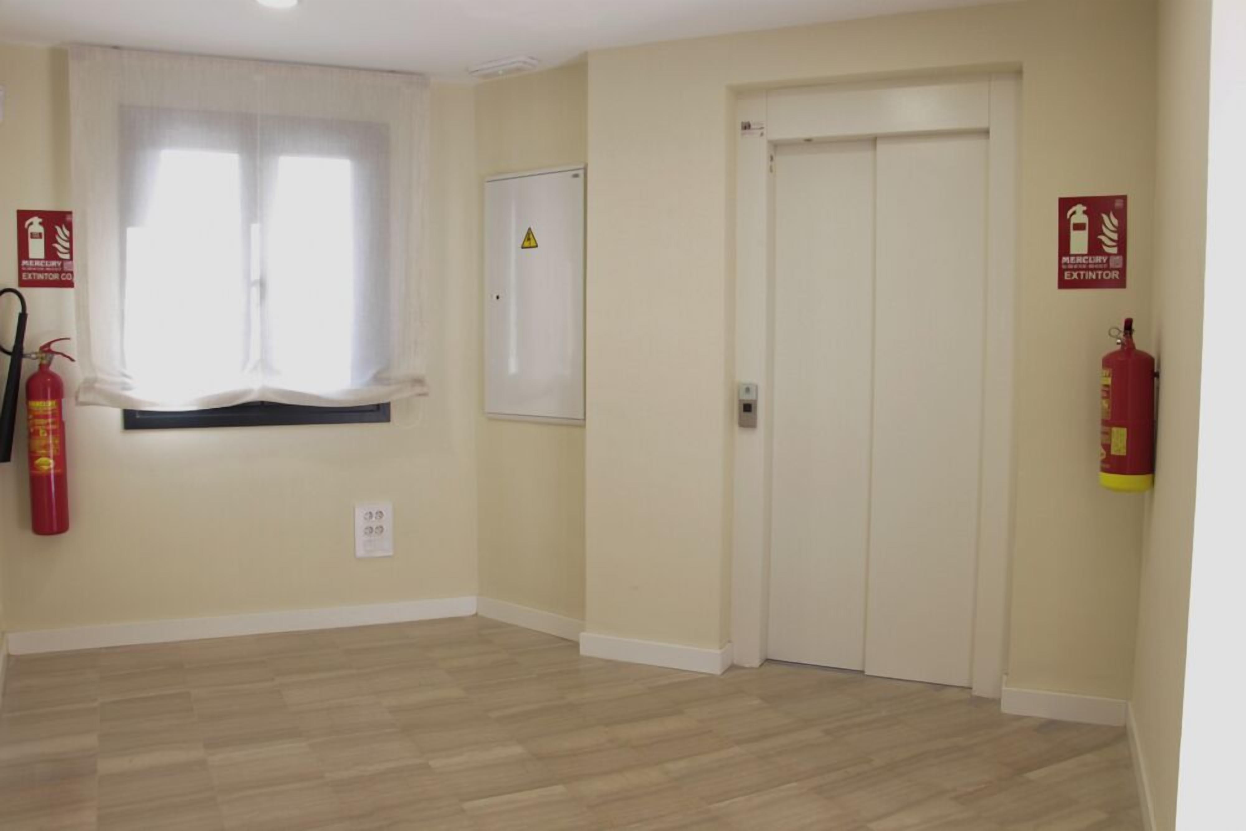 Una habitación con suelo de madera, una puerta blanca, un ventanal con cortinas translúcidas, una pizarra blanca y un extintor.