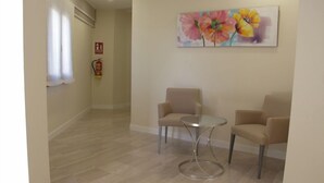Un couloir aux murs de couleur beige, avec un plancher en bois, deux fauteuils beiges, une petite table ronde et un tableau représentant des motifs floraux accroché au mur.