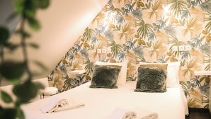 A hotel room with a bed, two pillows, a bedside table, and a wall with a tropical leaf pattern. - Maria Grazia Hotel (Saint-Gatien-des-Bois)