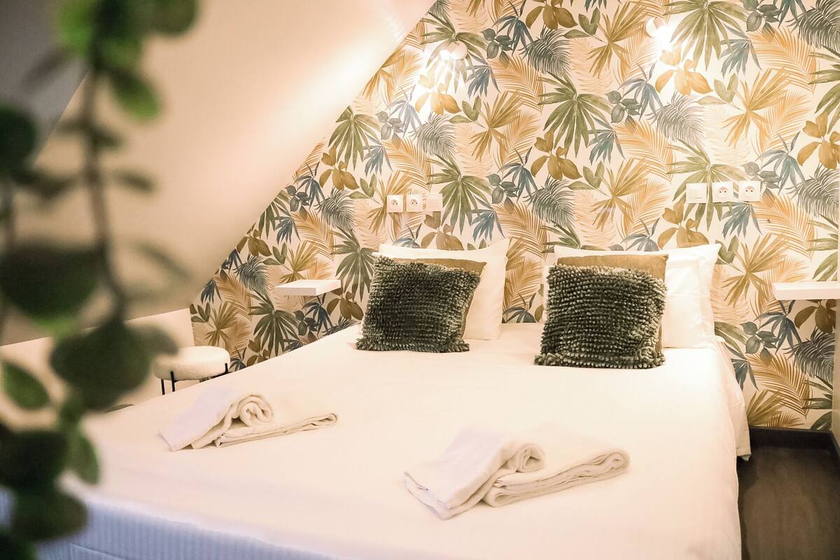 A hotel room with a bed, two pillows, a bedside table, and a wall with a tropical leaf pattern.