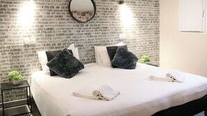 A hotel room with a brick-patterned wall, a large bed with white linens, two black textured pillows, and a hanging mirror. - Maria Grazia Hotel (Saint-Gatien-des-Bois)