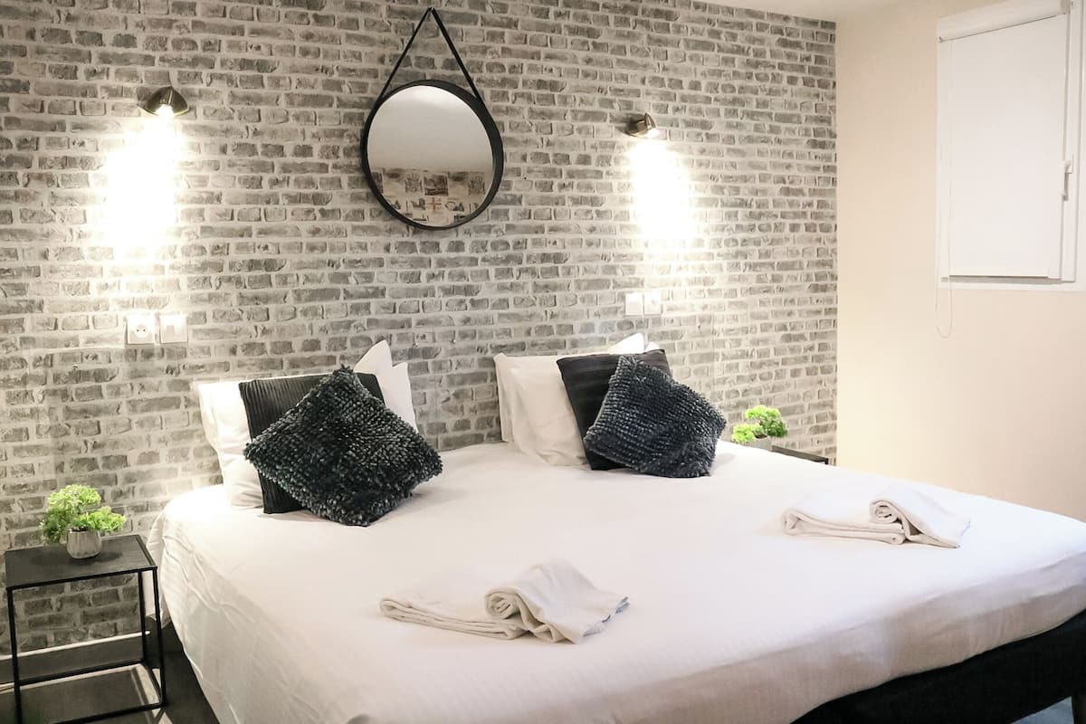 A hotel room with a brick-patterned wall, a large bed with white linens, two black textured pillows, and a hanging mirror.