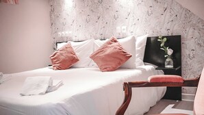 A neatly made bed with white linens and pink pillows, a wooden chair, and a wall with a tropical leaf pattern. - Maria Grazia Hotel (Saint-Gatien-des-Bois)