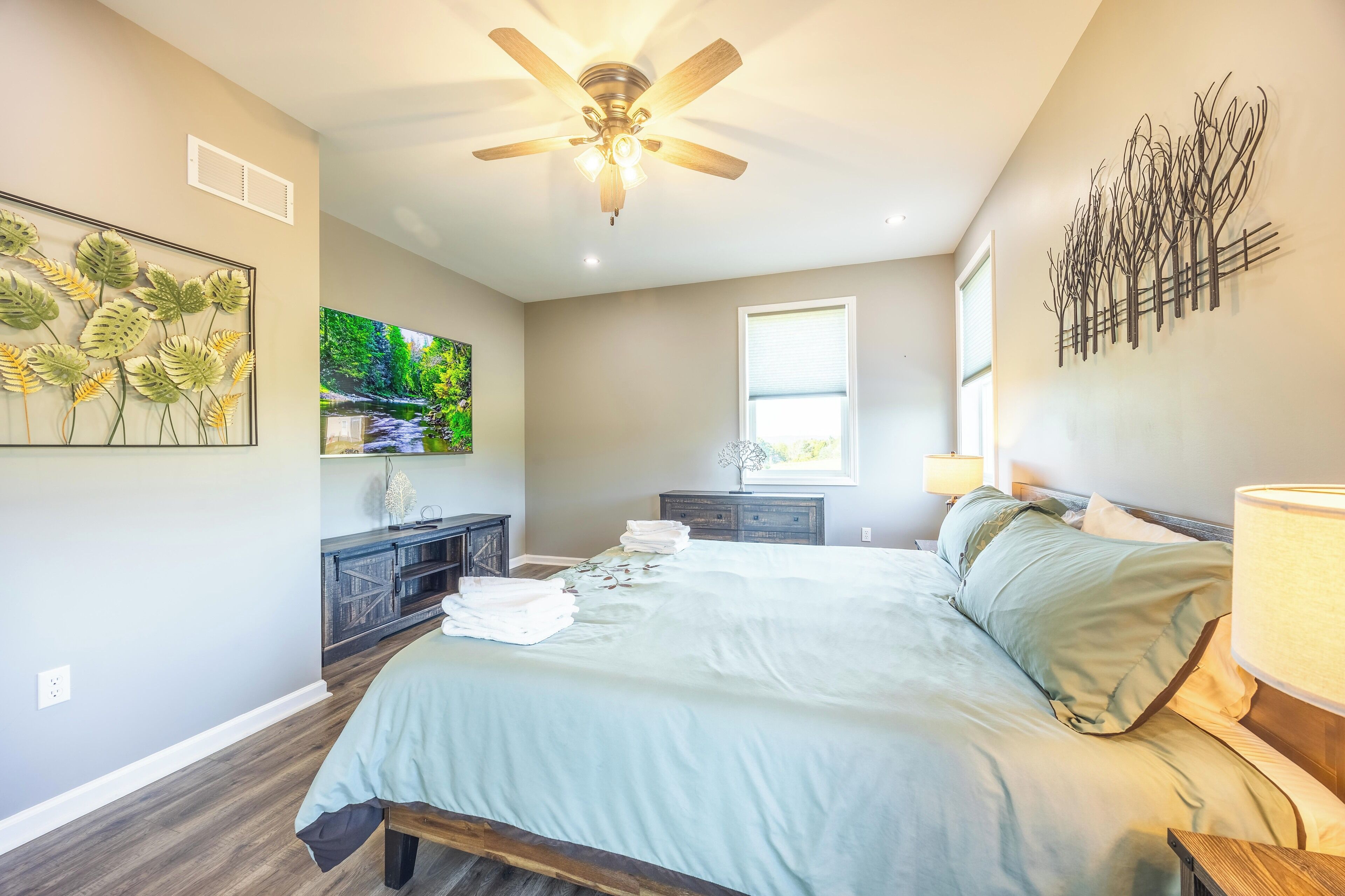 A bedroom with a bed, a ceiling fan, a TV, a window, and a wall art piece.