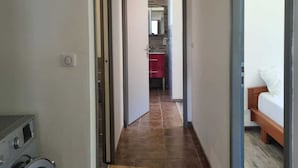 Un corridoio con lavatrice, un letto e una zona cucina visibile attraverso una porta aperta.