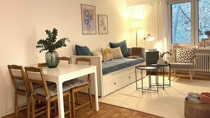 Una sala de estar acogedora con un sofá, una mesa de comedor con sillas, una mesita auxiliar y un ventanal con vistas a árboles.
