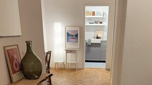Una habitación con piso de madera, una mesa blanca con un florero verde, un cuadro enmarcado, una silla blanca y una zona de cocina con armarios y un lavabo.