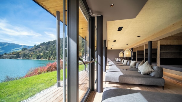 A modern interior with a large window overlooking a lake and hills, a wooden deck, and a seating area with sofas and lamps. - Hotel Seehof (Monguelfo)
