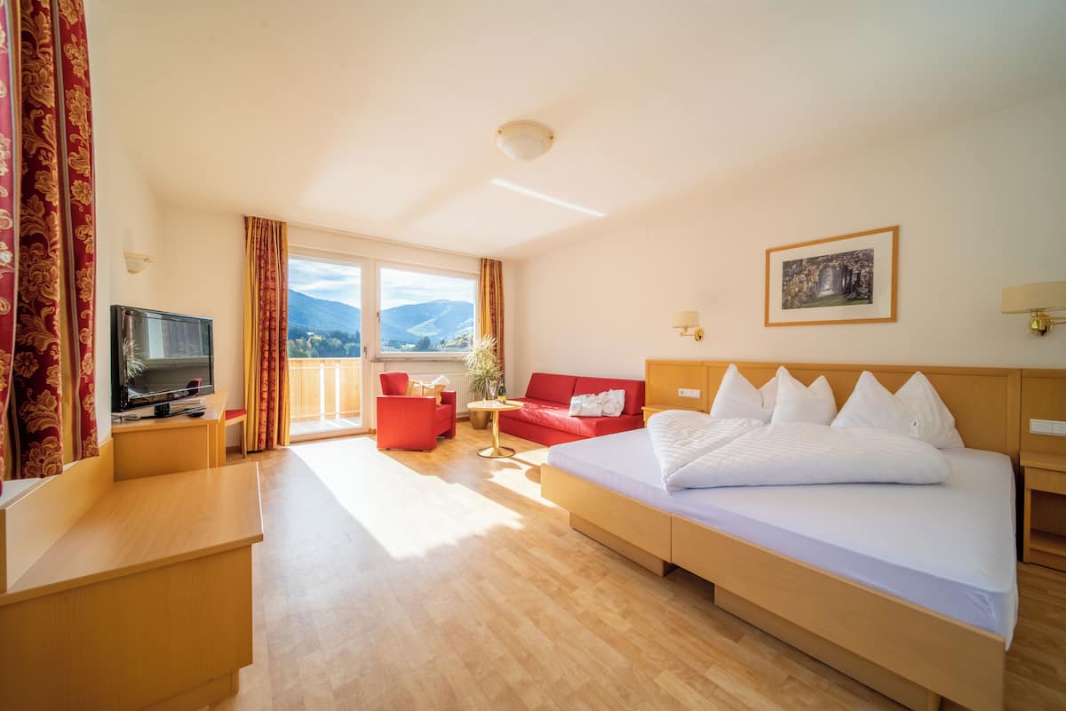 Une chambre d’hôtel avec un lit, un bureau, une télévision, un fauteuil rouge, un canapé et une fenêtre donnant sur la montagne.