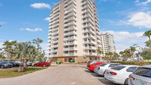 A high-rise apartment building with multiple balconies, a parking lot with several cars, and palm trees. - Island Getaway! Lovers Key Beach Club 604, Amazing Views And Private Beach! (Fort Myers Beach)
