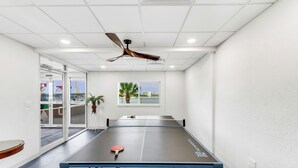 A ping pong table in a room with a ceiling fan, a window with a view, and a door leading outside. - Island Getaway! Lovers Key Beach Club 604, Amazing Views And Private Beach! (Fort Myers Beach)