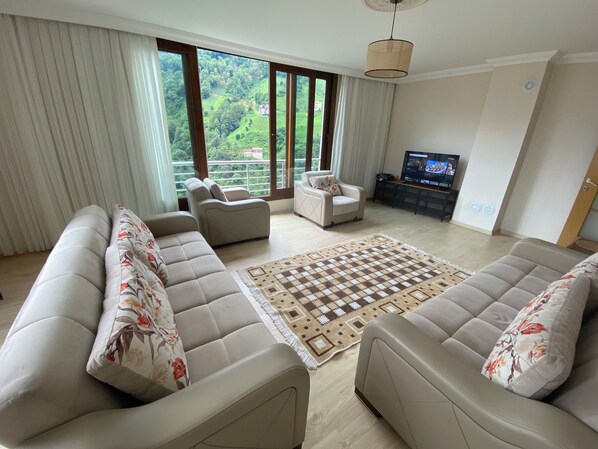A living room with a sofa set, a television, and a large window with a view of greenery.