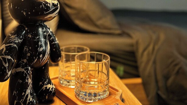 A bear figurine on a wooden tray with two glasses, a lamp, and a bed with pillows in the background. - FlexiHome (Ho Chi Minh City)