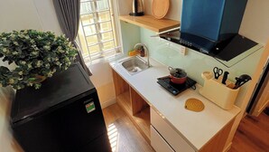 A compact kitchen with a built-in stove, a sink, and a small countertop. There is a window with curtains, a potted plant, and a shelf with kitchen utensils. - FlexiHome (Ho Chi Minh City)