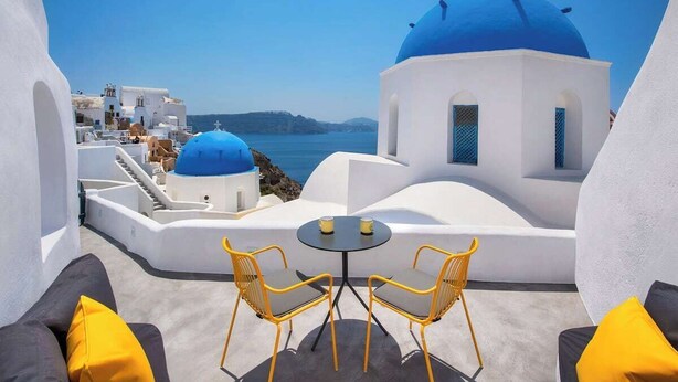 A terrace with white buildings, blue domes, and a view of the sea.