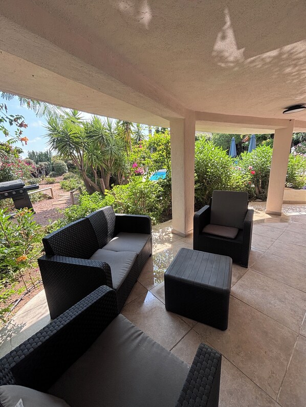 A covered outdoor seating area with wicker furniture, a small table, and a view of a garden with various plants and a blue umbrella.
