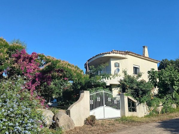 A two-story house with a curved roof and a chimney, surrounded by lush greenery and flowering plants.