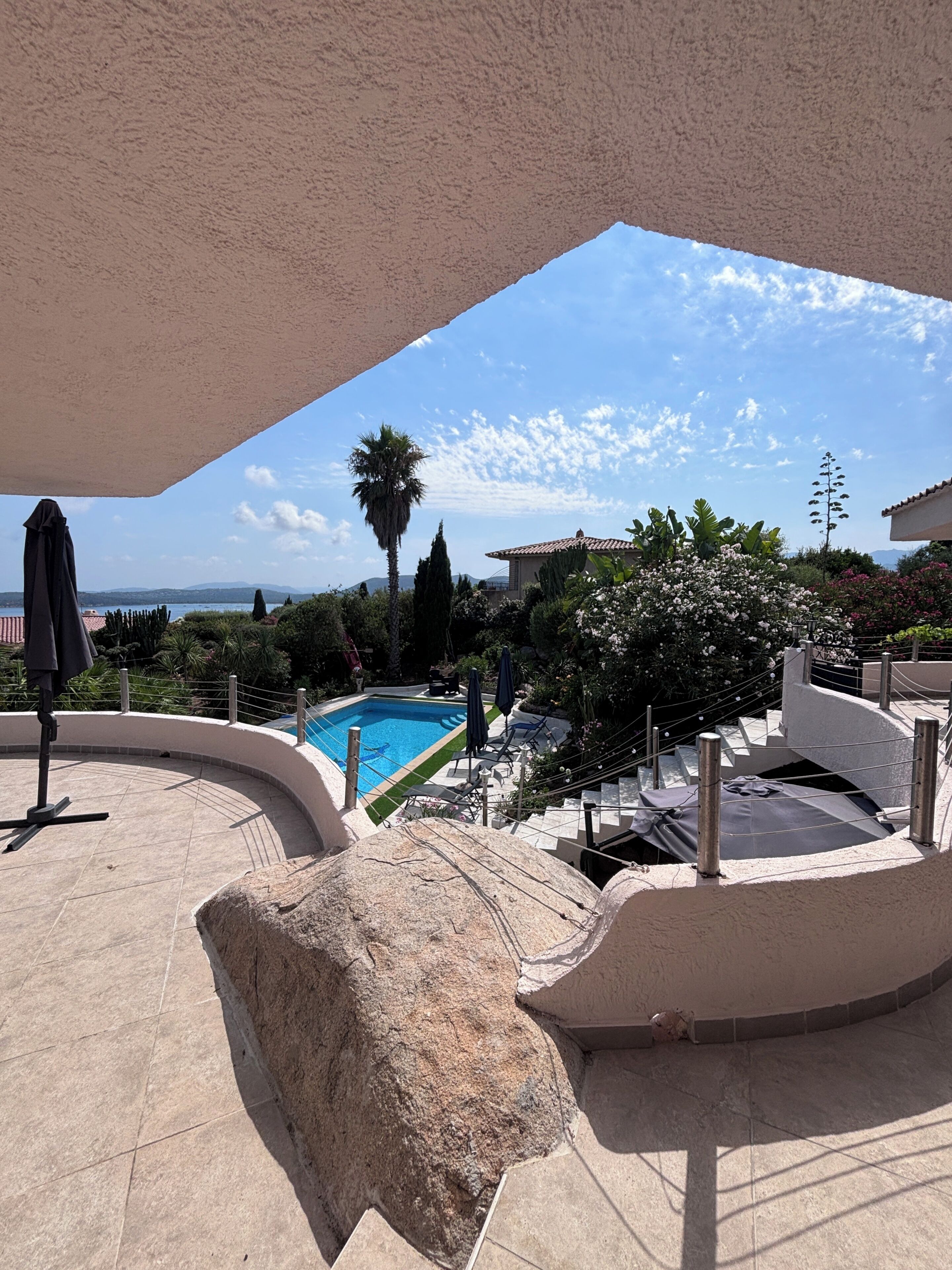 A swimming pool with a view of the sea, surrounded by greenery and a clear sky.