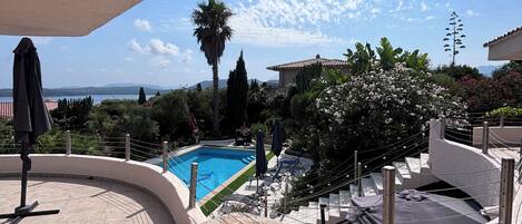 A swimming pool with a view of the sea, surrounded by greenery and a clear sky.