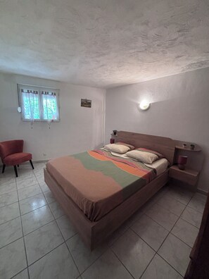 A bedroom with a bed, a chair, a nightstand, a wall-mounted light, and a window with curtains.