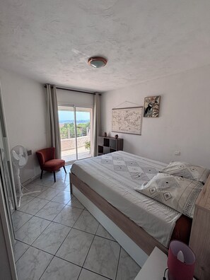 A bedroom with a bed, a chair, a fan, a map on the wall, and a view through a window.