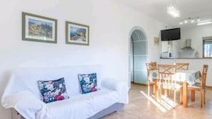 Een moderne woonkamer met een witte bank, bloemenkussens, een eethoek met houten stoelen en een keuken op de achtergrond.