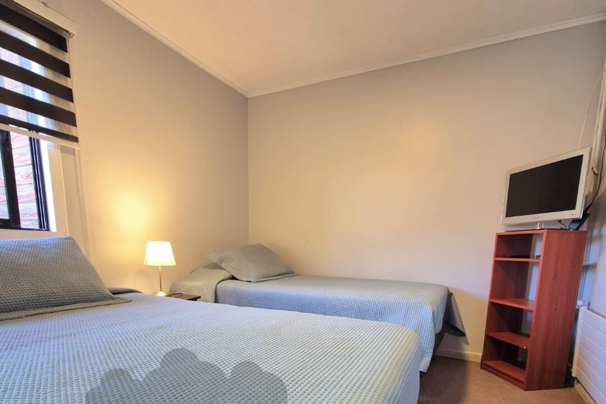 Ein kleines, sauberes Zimmer mit zwei Betten, einem Fernseher auf einem Holzschrank und einem Fenster mit Jalousien.