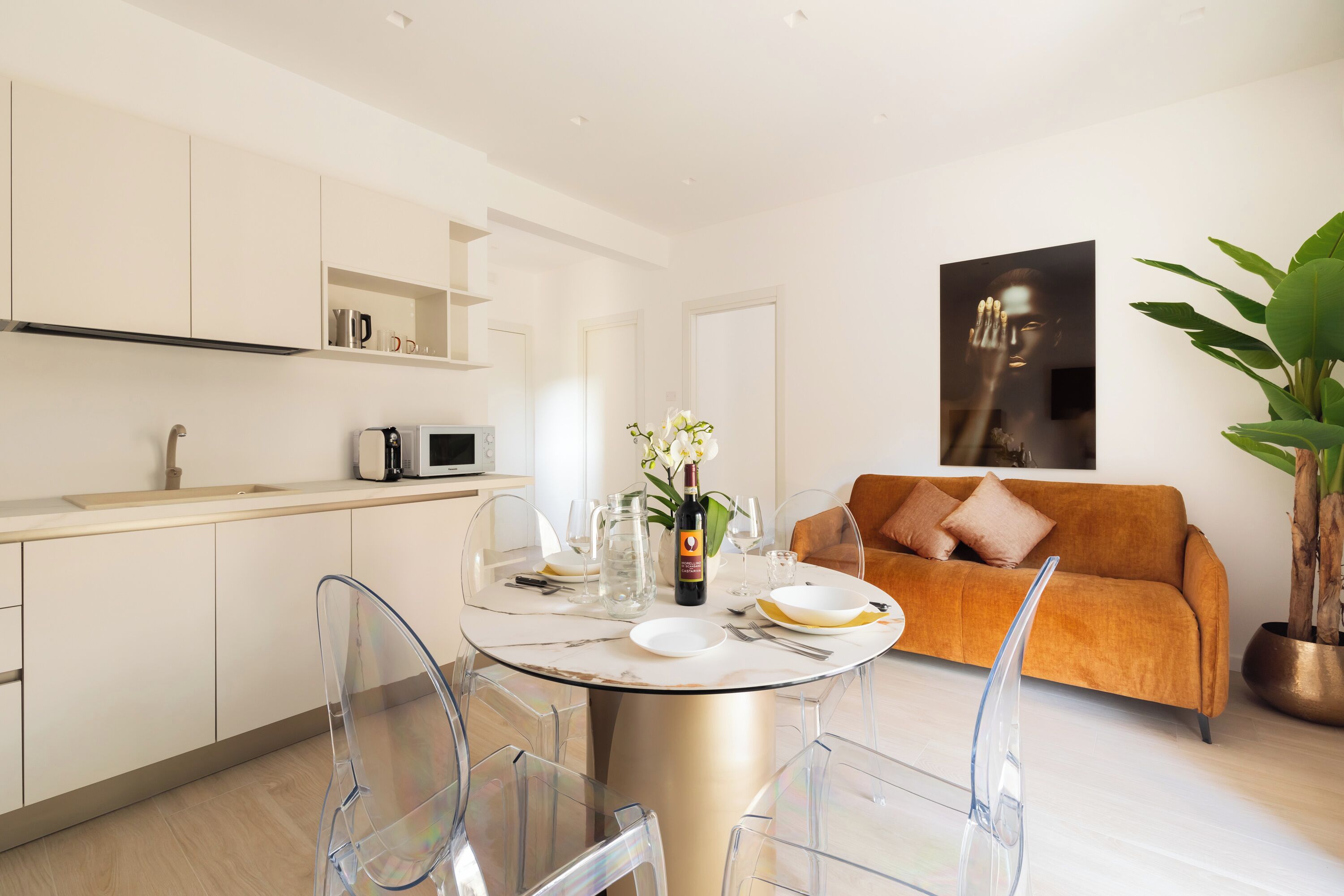 A modern kitchen with white cabinets, a dining table with clear chairs, and a living area with a sofa and a plant.
