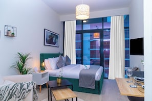 A modern hotel room with a bed, bedside table, lamp, and a view of the cityscape through large windows.