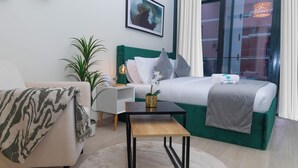A modern hotel room with a green bed, white pillows, a small wooden table, a white armchair, and a large window with curtains. - Bloomfields Fantastic STU Azizi Riviera (Dubai)