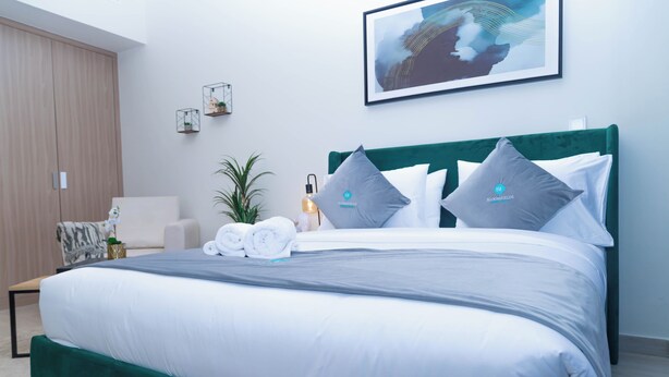A neatly made bed with white linens and two grey pillows, a green upholstered headboard, and a wall-mounted abstract painting. - Bloomfields Fantastic STU Azizi Riviera (Dubai)