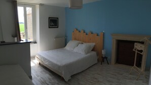 Un dormitorio con cabecera de madera, cama blanca, mesita de noche pequeña y chimenea, además de una ventana con cortinas.