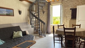 Una sala de estar acogedora con un sofá, una mesa de centro de madera, una mesa de comedor con sillas, una escalera de caracol y una ventana con vistas a la vegetación.